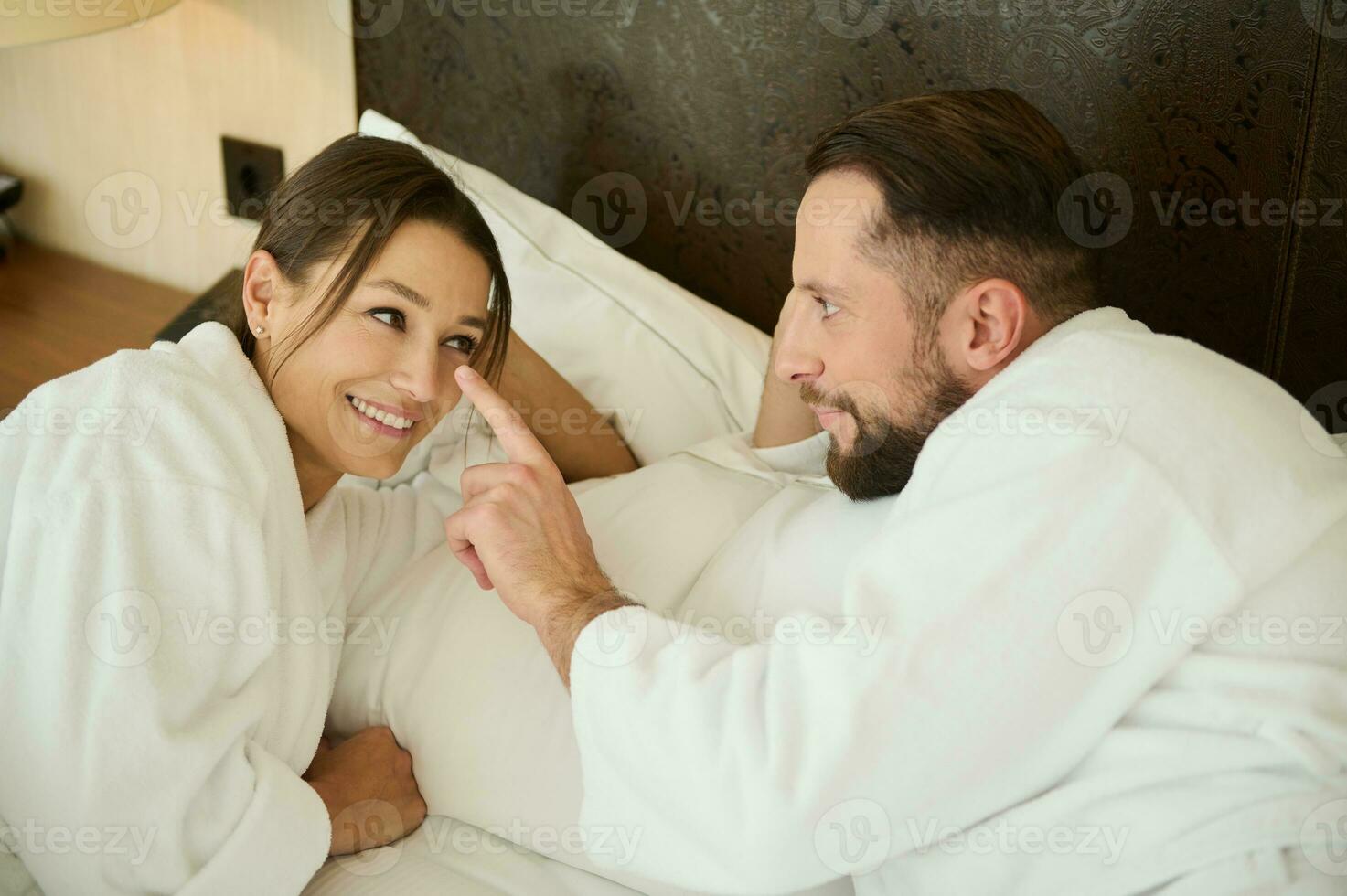 un hermoso joven hombre, amoroso marido suavemente conmovedor su amado de la esposa nariz con su dedo, tendido siguiente a su en cama. joven contento Pareja en amar, disfrutando unión en su Luna de miel. foto