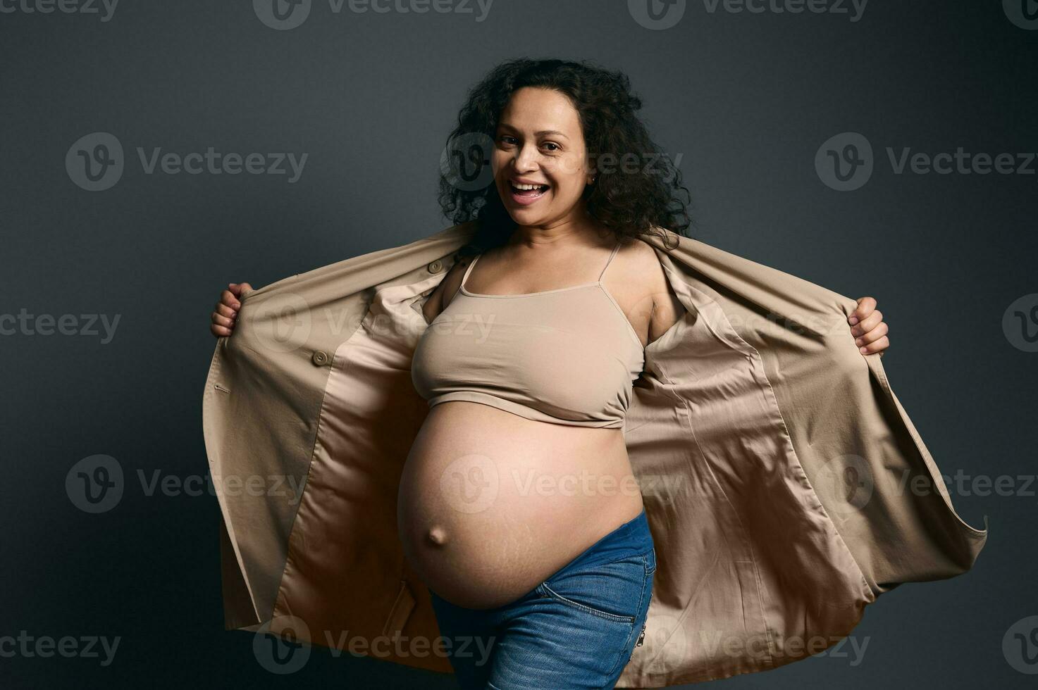 contento Rizado embarazada mujer en tarde el embarazo, demostración su desnudo barriga, sonriente terminado gris estudio antecedentes. foto