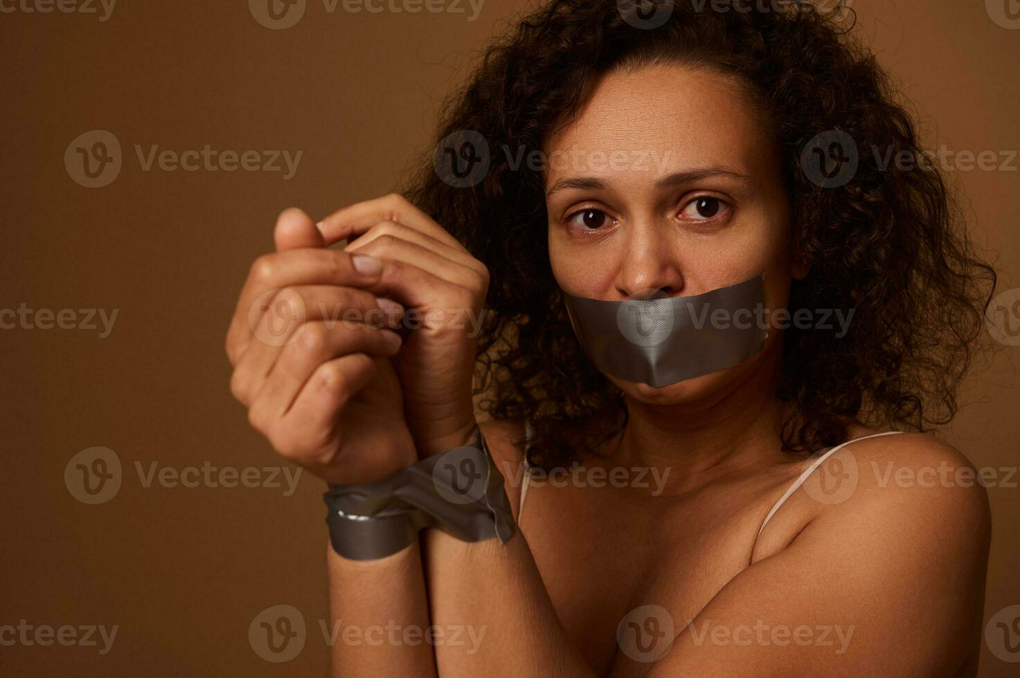 Close-up portrait of a scared half naked mixed race woman with tied hands and closed mouth, looking at camera, isolated on colored dark background with copy space. Elimination violence against women. photo