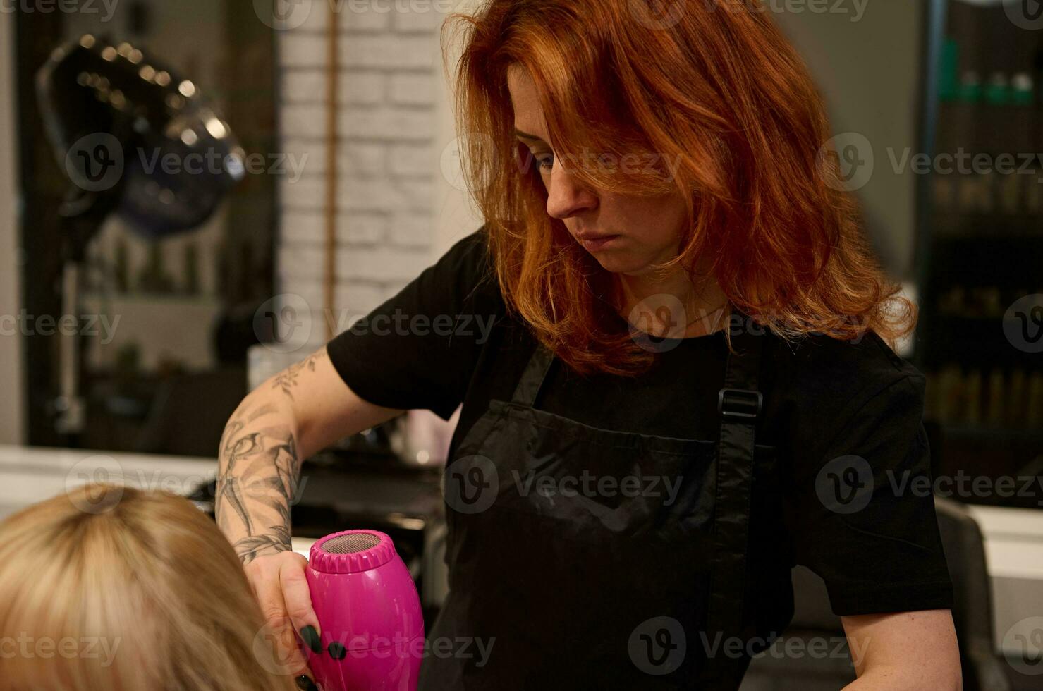 de cerca de un de edad mediana pelirrojo mujer peluquero con tatuado brazos en un estricto negro uniforme peinada, enderezamiento y el secado el rubia pelo de un cliente de un belleza salón utilizando un rosado secador de pelo foto