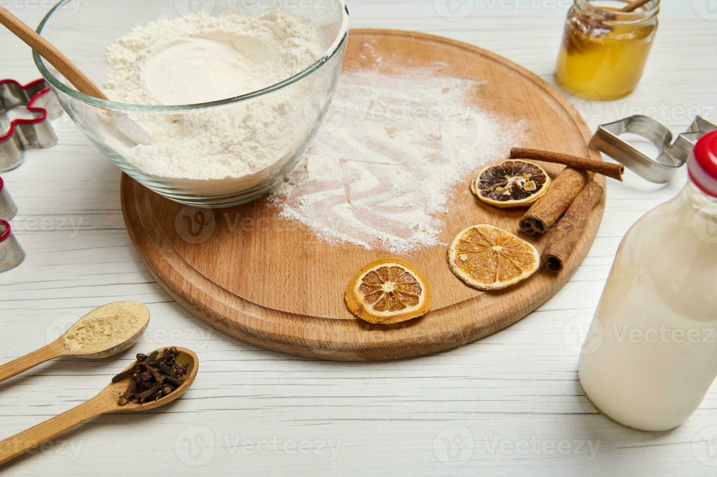 de cerca de ingredientes para preparando masa y pan de jengibre galletas, corte moldes, botella con planta establecido leche, vaso tarro con miel y un cuenco con harina en un redondo de madera tablero foto