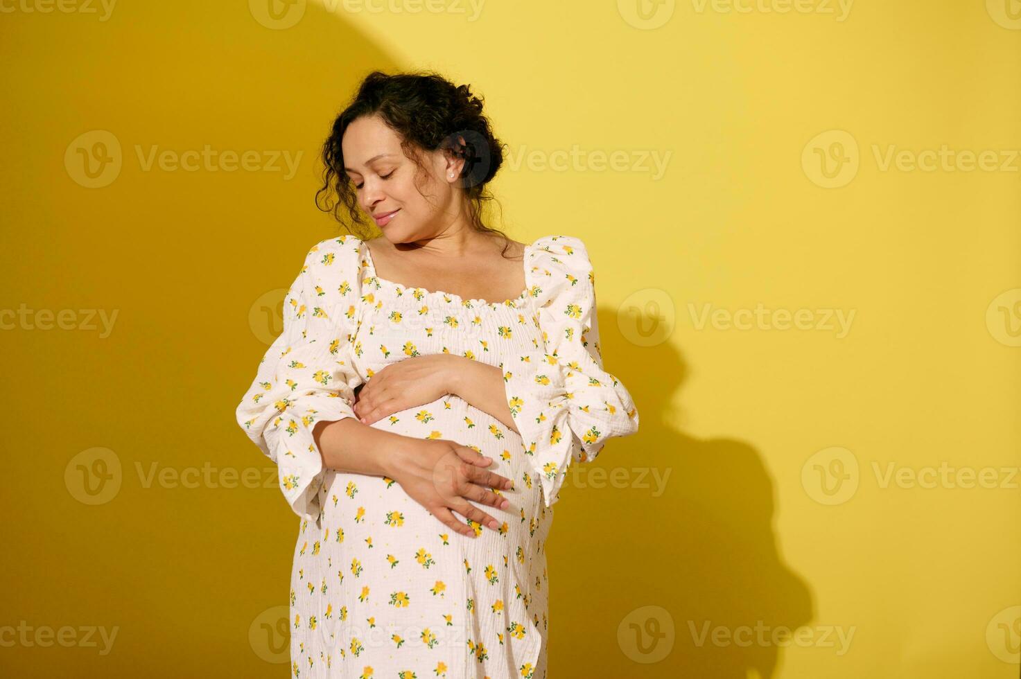 Delightful pregnant woman gently stroking and hugging her big belly, isolated over yellow studio background photo