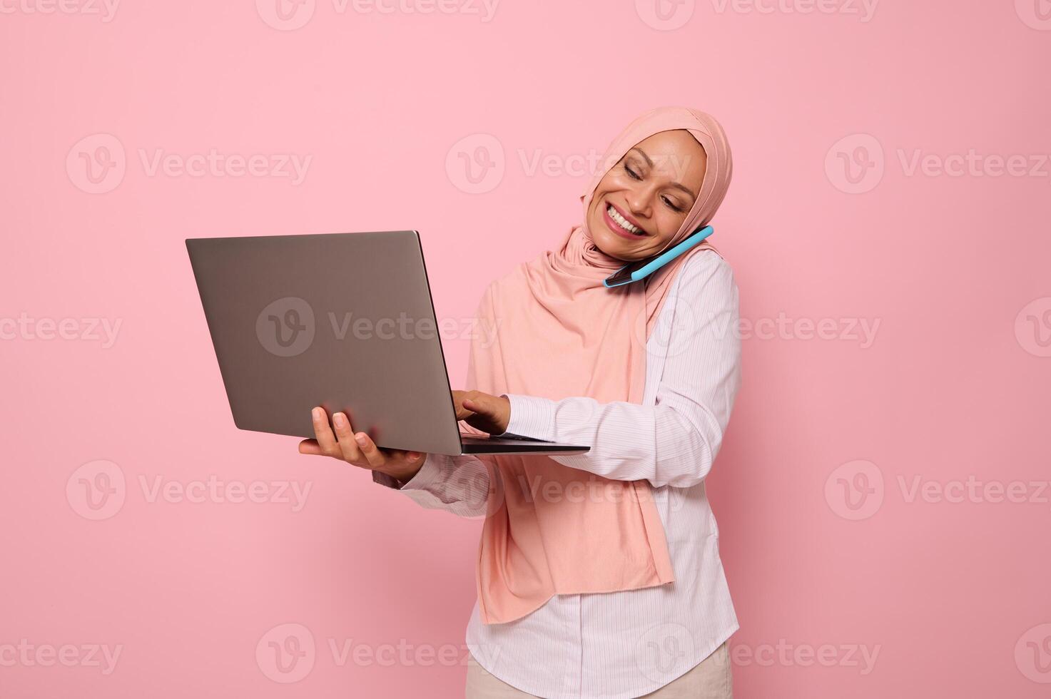 Successful Muslim Arab beautiful woman in hijab, dressed business smart casual talks on mobile phone , smiles looking at the monitor screen while typing text on a laptop. Copy space, pink background photo