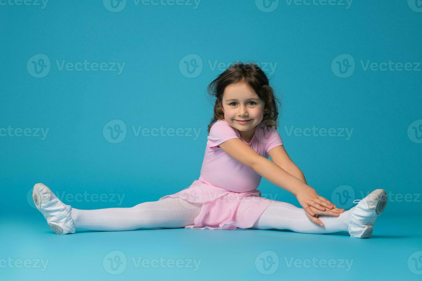 hermosa pequeño niña bailarina sentado en un azul fondo, practicando y extensión su cuerpo foto