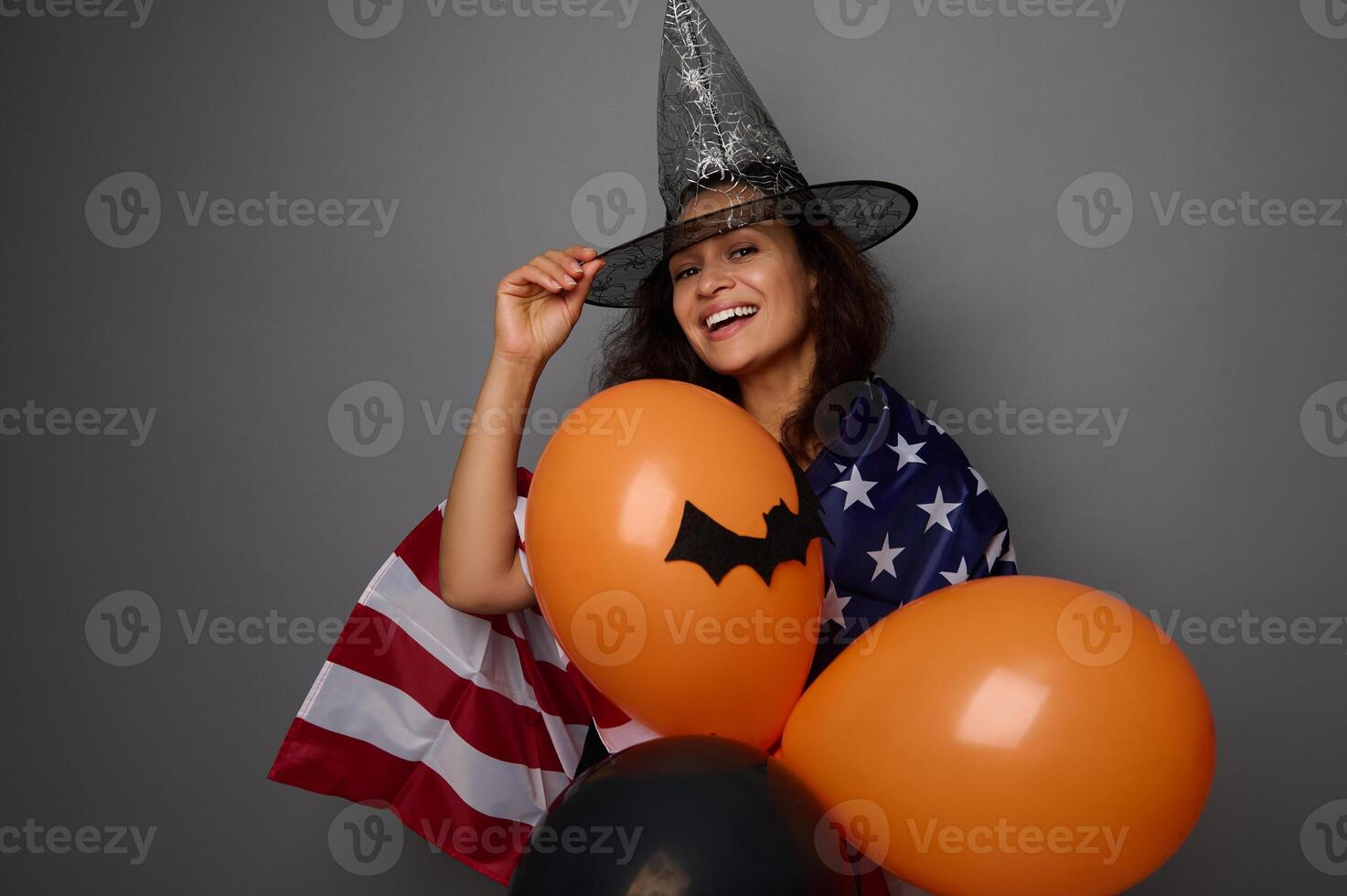 Cheerful mixed race young woman in wizard hat, wrapped in American flag, holds colorful orange air balls, smiles toothy smile looking at camera. Halloween concept on gray background with copy space photo