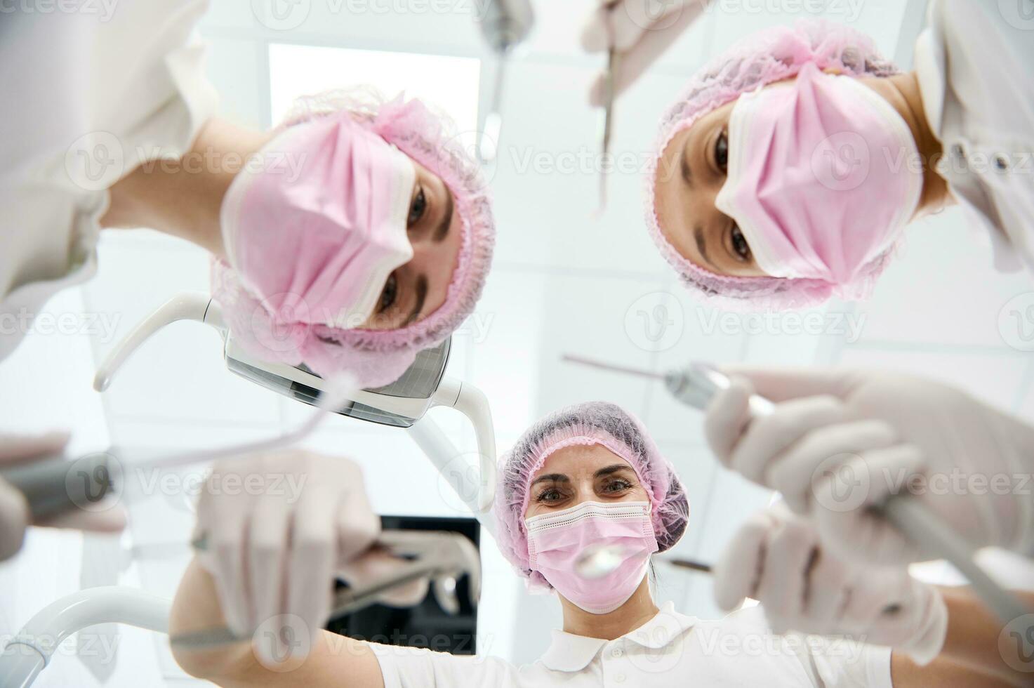 fondo ver de hembra dentistas y dental asistente en rosado protector máscara participación estéril herramientas para un dental examen y propensión hacia un paciente . atención en dental instrumentos foto