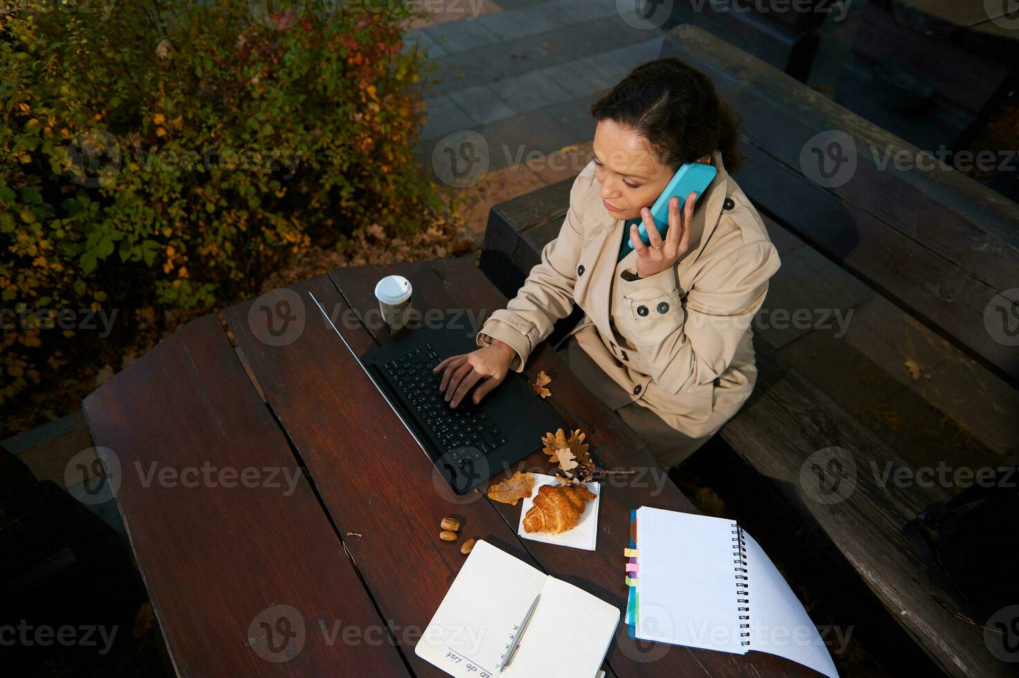 parte superior ver de un negocio mujer, confidente persona de libre dedicación, hablando en móvil teléfono, mecanografía en ordenador portátil teclado, disfrutando distante trabajo en un de madera café en un roble arboleda en hermosa otoño calentar soleado día foto