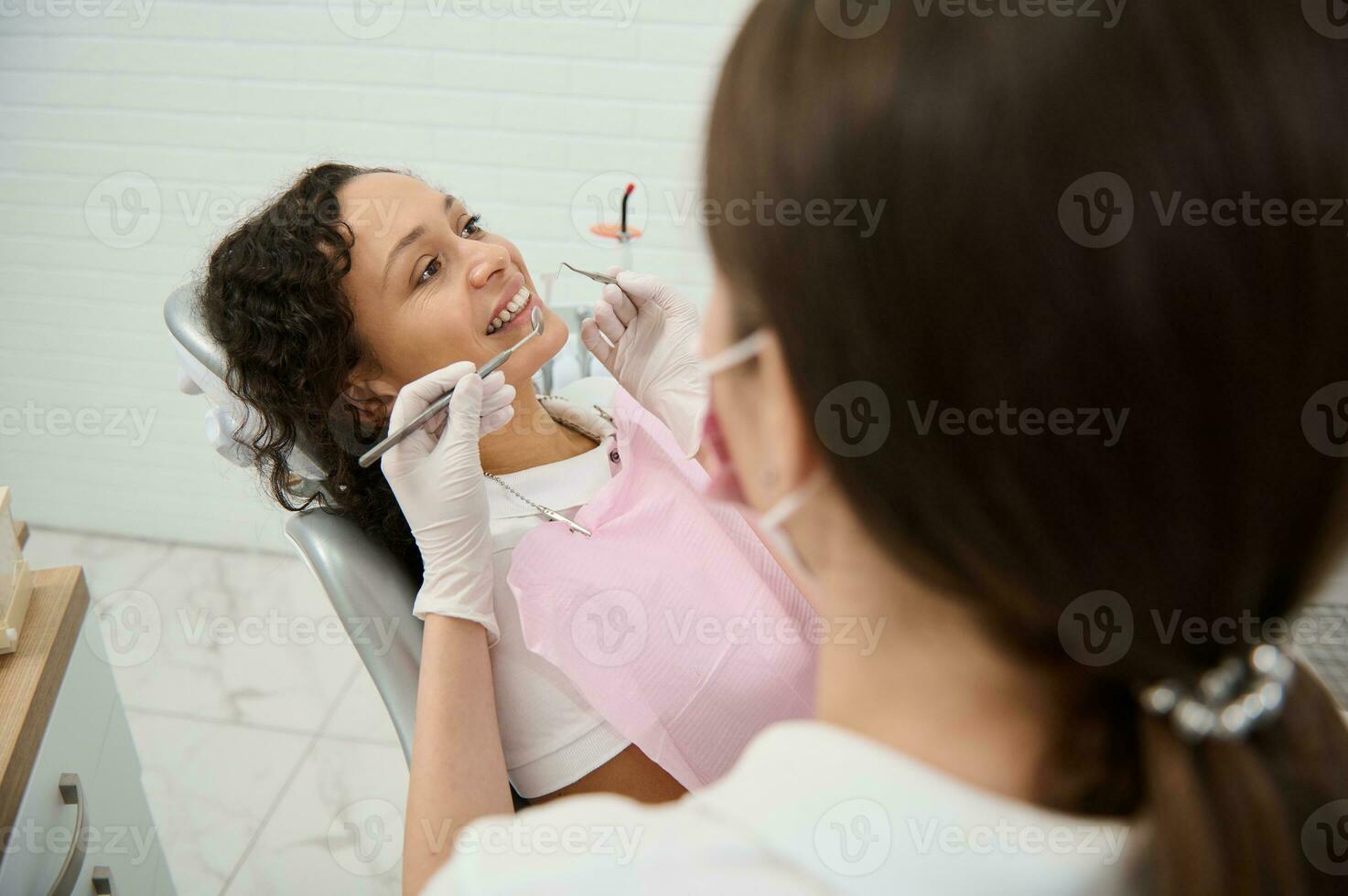View through blurred dentist hygienist holding stainless steel dental tools- probe, mirror and examining the oral cavity of a dentistry clinic patient with perfect white teeth sitting in dentist chair photo