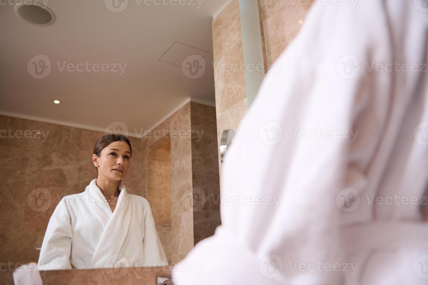 Mirror reflection of an attractive confident middle aged brunette woman wearing bathrobe looking at herself standing in bathroom during morning hygiene routine photo