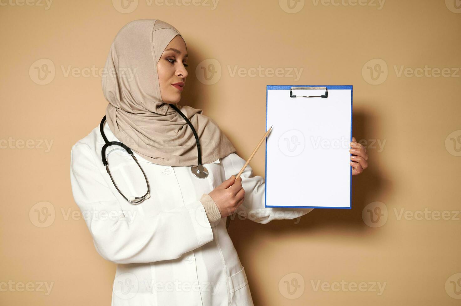 Serene and handsome doctor in hijab holding a clipboard with a blank sheet of paper and pointing at it. Shot with soft shadow. Beige background with copy space. photo