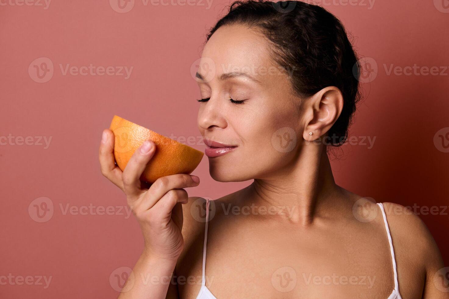 de cerca de pelinegro bonito mujer oliendo el olor de Fresco jugoso rojo pomelo, posando con cerrado ojos en contra coral antecedentes con Copiar espacio. salud, cuerpo positividad y piel cuidado concepto. foto