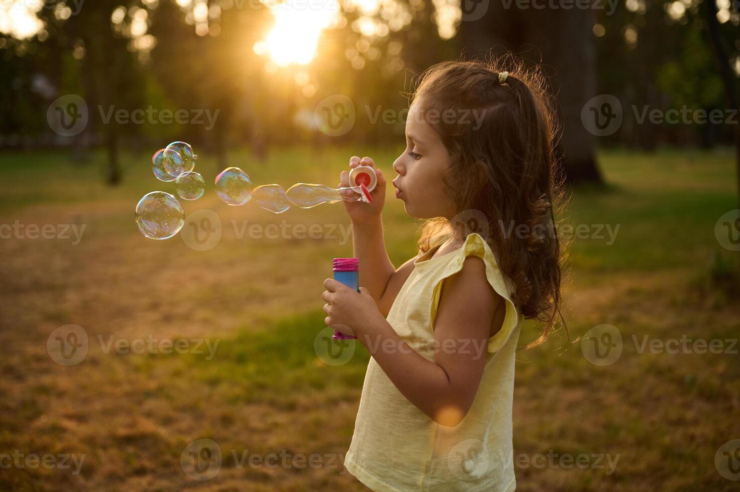 Adorable child baby girl, blowing soap bubbles at sunset, enjoying pleasant time outdoors in the meadow. Sun's rays fall through transparent bubble spheres with iridescent reflections. photo