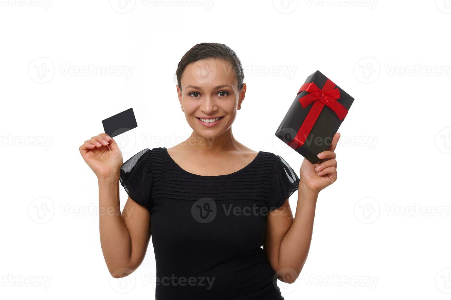 Isolated portrait on white background with copy space of beautiful smiling African American woman dressed in black, holding discount credit card and gift box, looking at camera. Black Friday concept photo