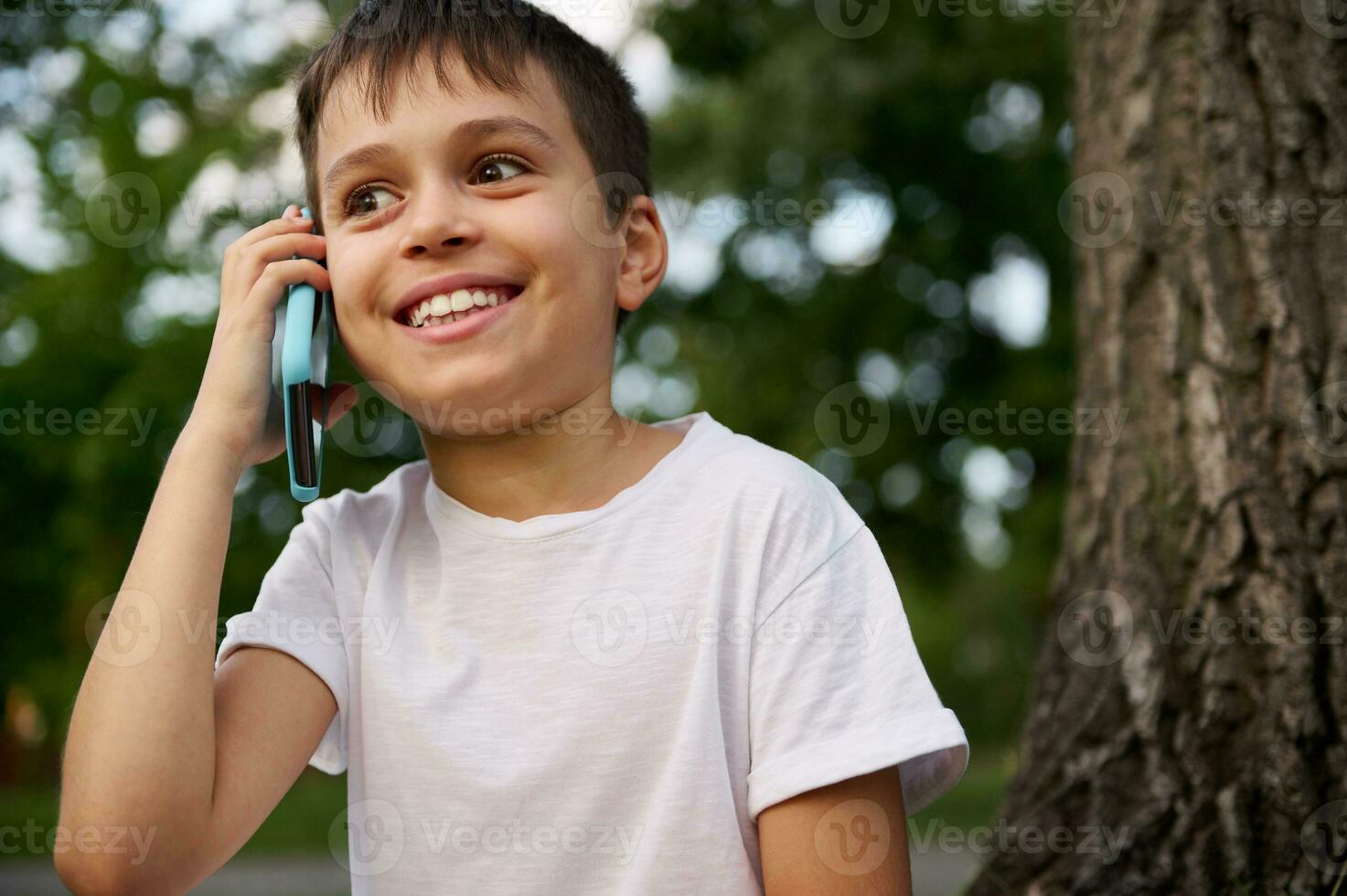 Cheerful adorable school child boy talking on mobile phone, cute smiling looking away, resting on the public park after first day at school on beautiful summer day. Concepts of Back to school photo