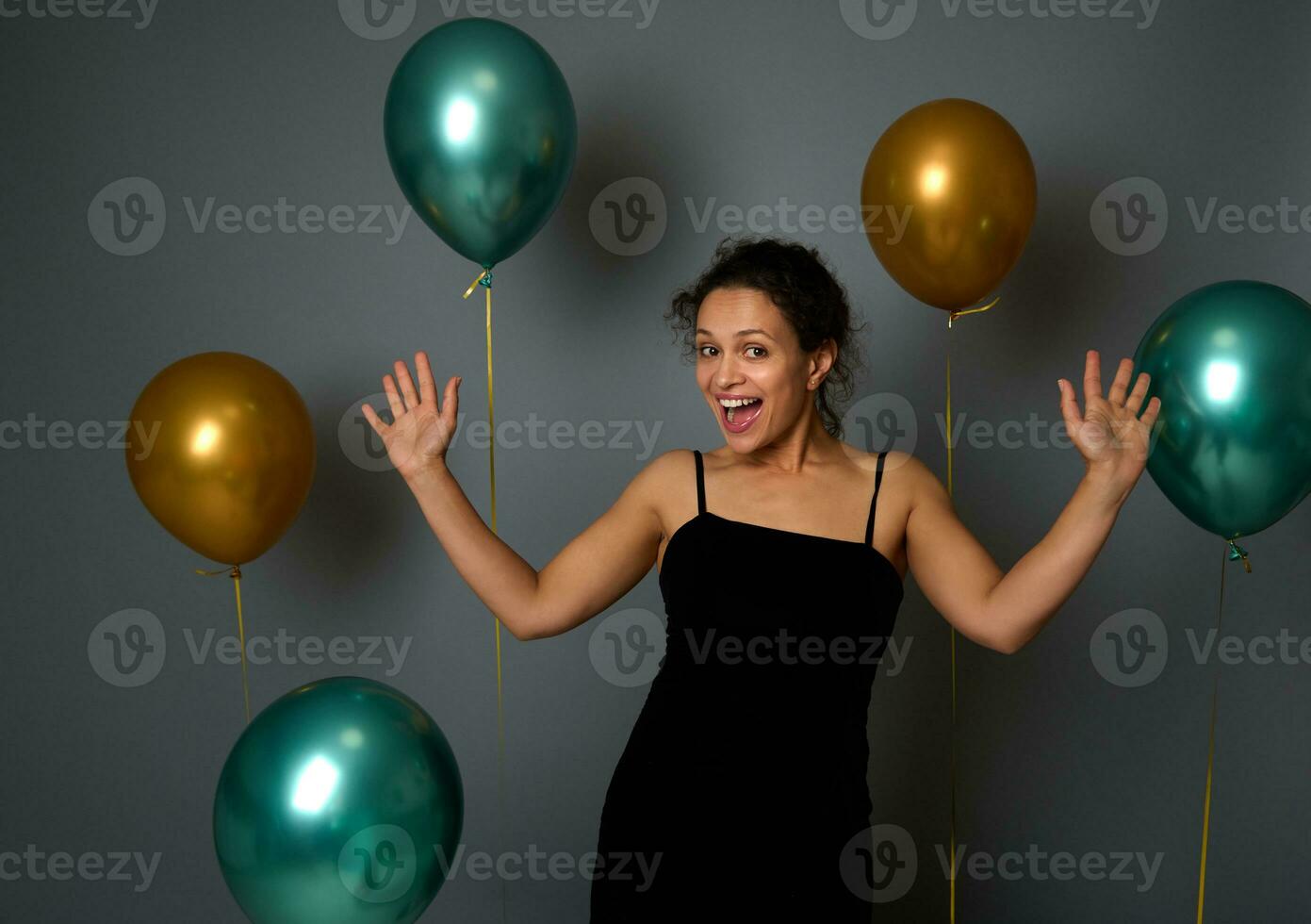 Cheerful young Latin woman in black evening dress rejoices, smiles toothy smile looking at camera against a festive ornate gray wall background with beautiful shiny gold and green metallic balloons photo