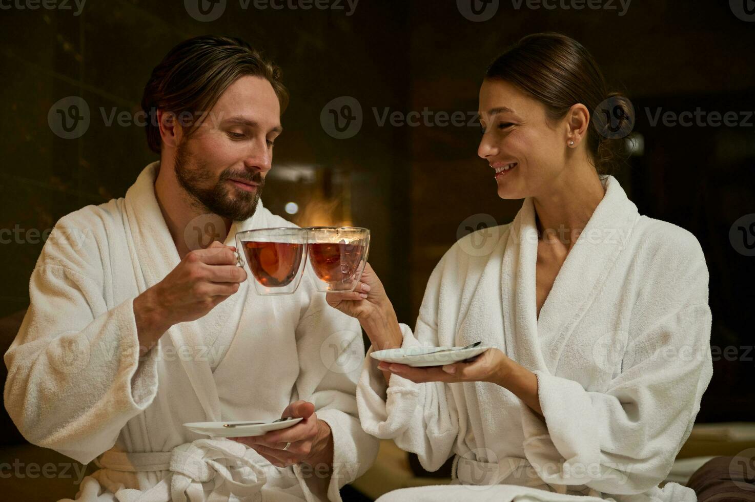 Happy newlywed, charming middle aged married Caucasian couple in white bathrobes holding cups with herbal tea, sitting close to each other, face to face, enjoying time together in wellness spa resort photo