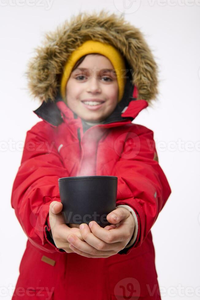 Adorable cheerful school-age boy in bright warm clothes smiles toothy smile, looking at the camera, warming his hands on a mug with a hot tea drink. Winter vacation travel concept with copy ad space photo