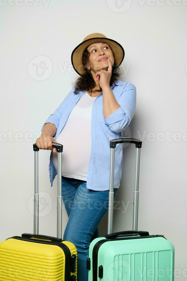 Happy multiethnic gravid woman dreamily looking away, standing with yellow and blue suitcases on isolated background photo