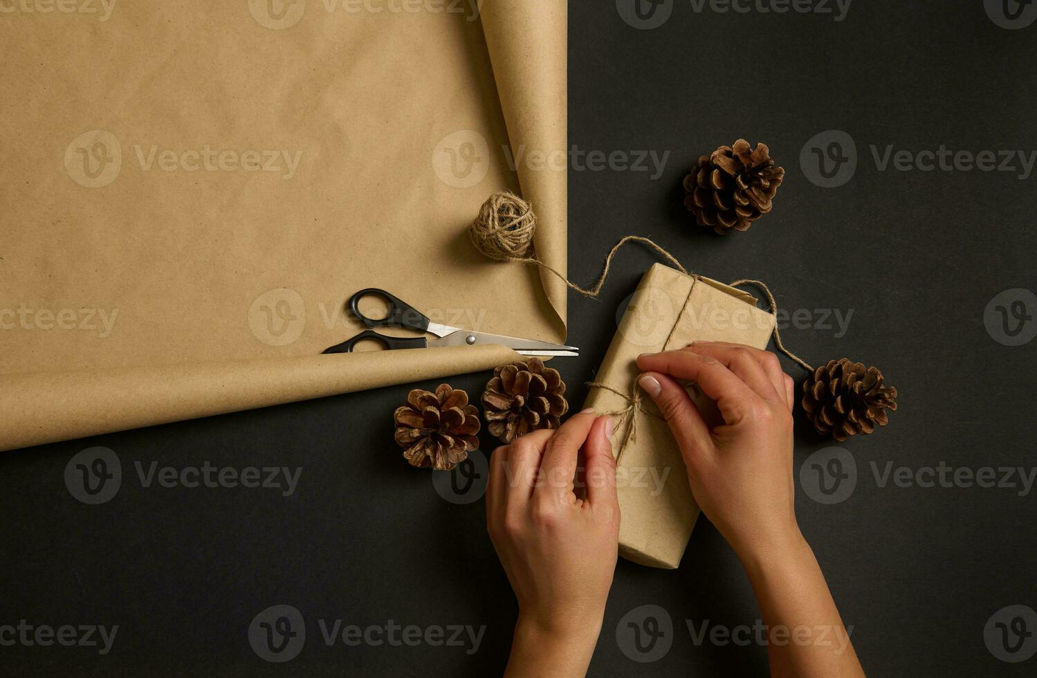 High angle view of female hands wrapping Christmas present in craft paper tying bow with rope on black surface with brown cardboard paper and scissors. Christmas traditional handmade decoration photo