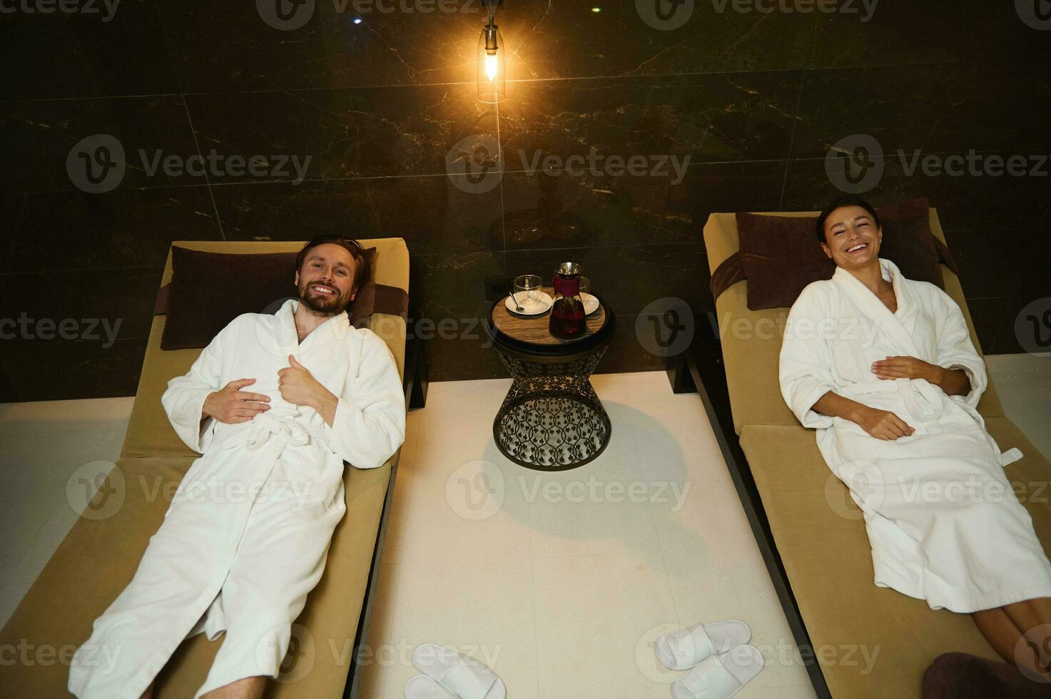 Relaxed happy loving young married couple smiles toothy smile looking at camera while resting in white terry bathrobes lying down on chaise lounges in lounge area of a luxury wellness spa complex photo