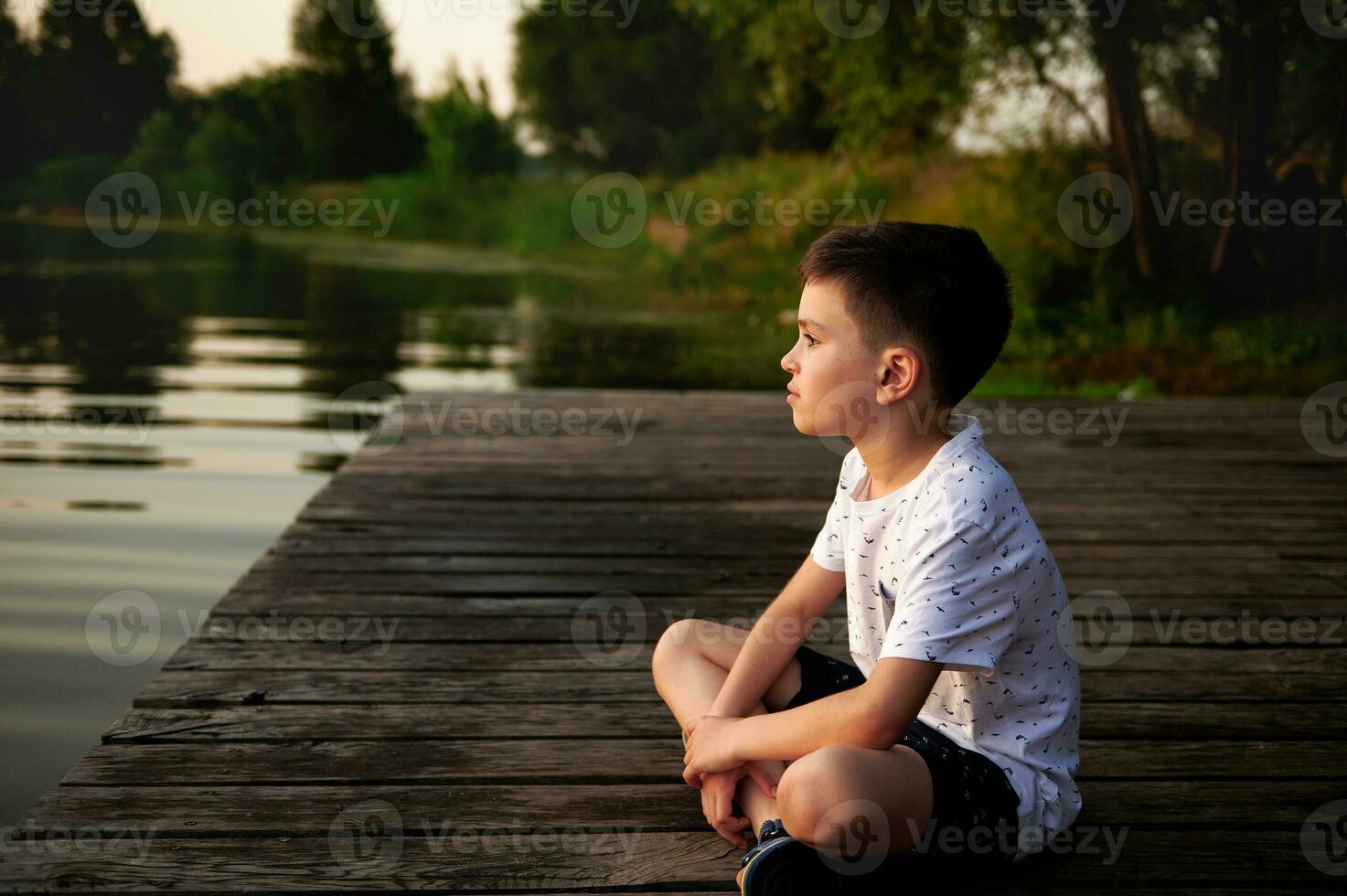 A charming boy resting on the pier and looking into the distance against the backdrop of the river bank. Camping, summer themes. photo