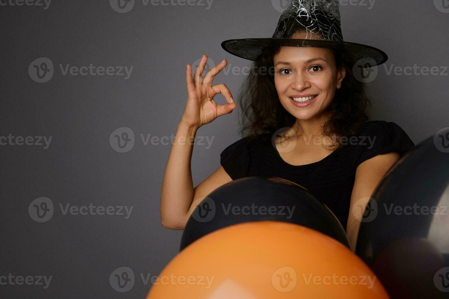 Close-up of beautiful smiling Hispanic woman in wizard hat, dressed in witch carnival costume for Halloween party, shows OK sign, poses against gray background with black balloons, copy space photo