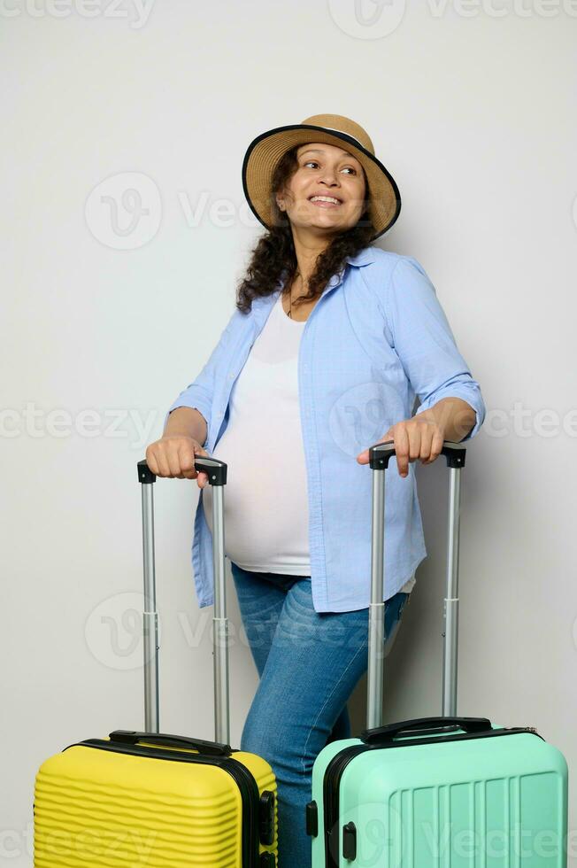 Beautiful pregnant woman travelling abroad, smiles looking aside, poses with suitcases, isolated white studio background photo