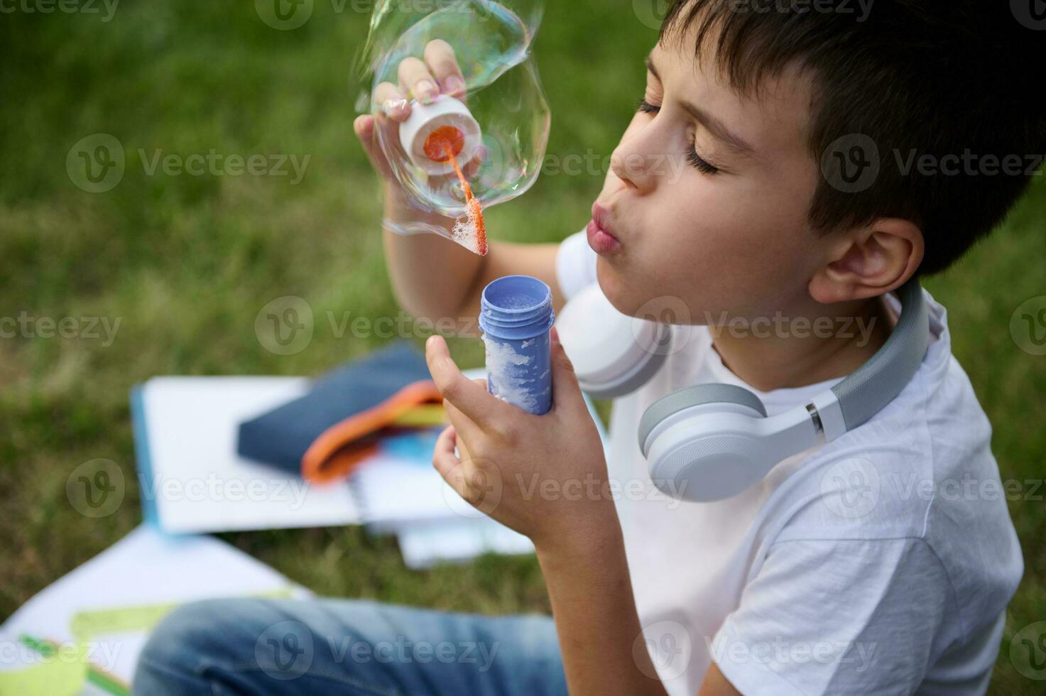 Headshot of an elementary aged schoolboy resting in the park sitting on the green grass after school, blowing bubbles. School bag with workbooks and school supplies lying down on the grass photo