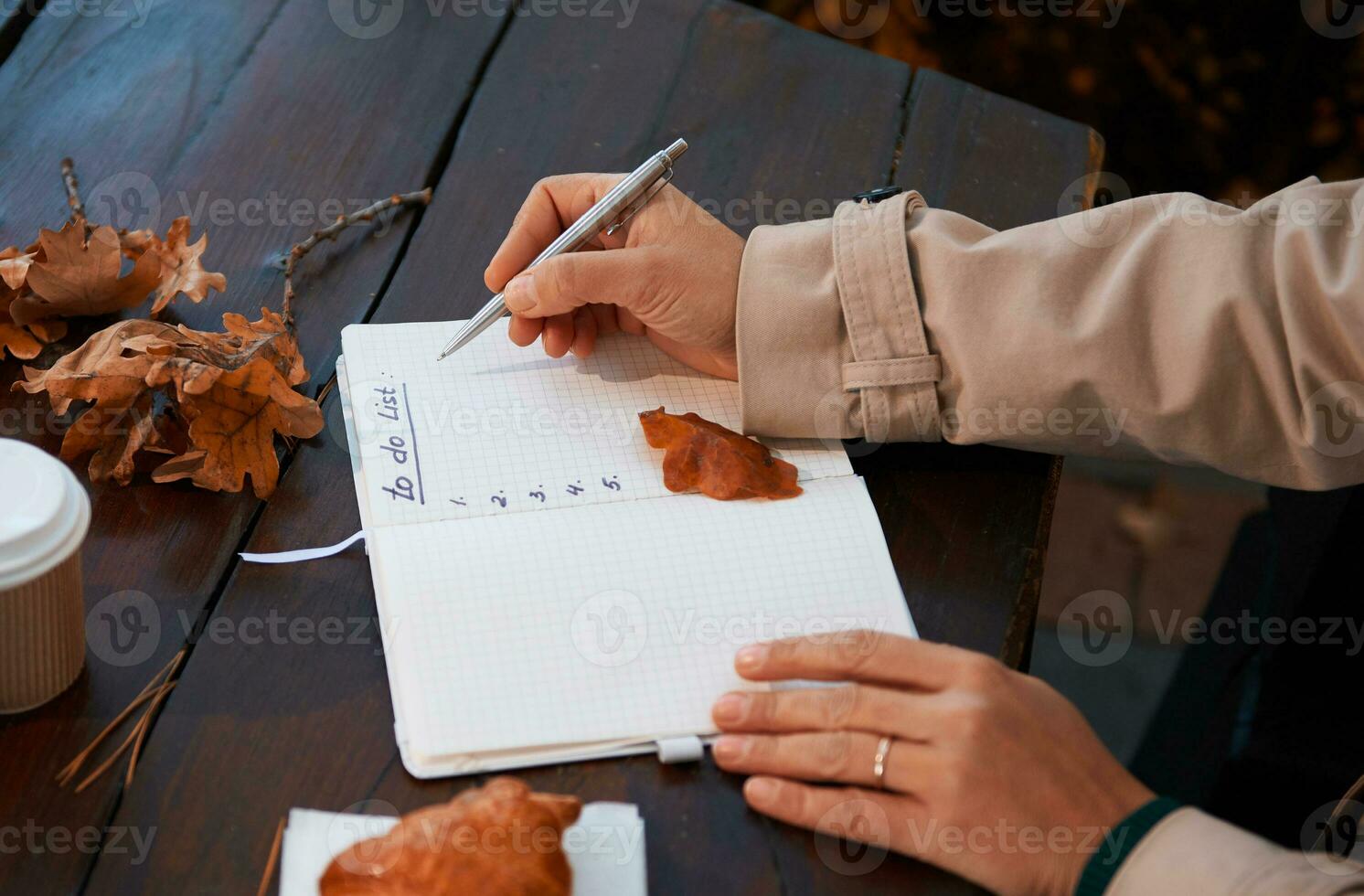 suave atención en hembra mano participación plata bolígrafo y escritura el lista a hacer en bloc, mientras sentado a un de madera mesa con caído seco roble hojas, para llevar papel taza y horneado cuerno foto