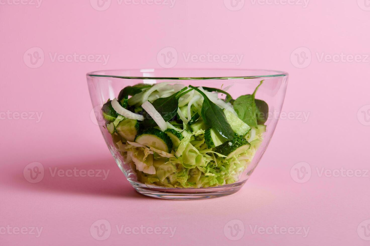 Close up of green salad with herbs, lettuce, cabbage and cucumber in transparent glass bowl on pink background photo