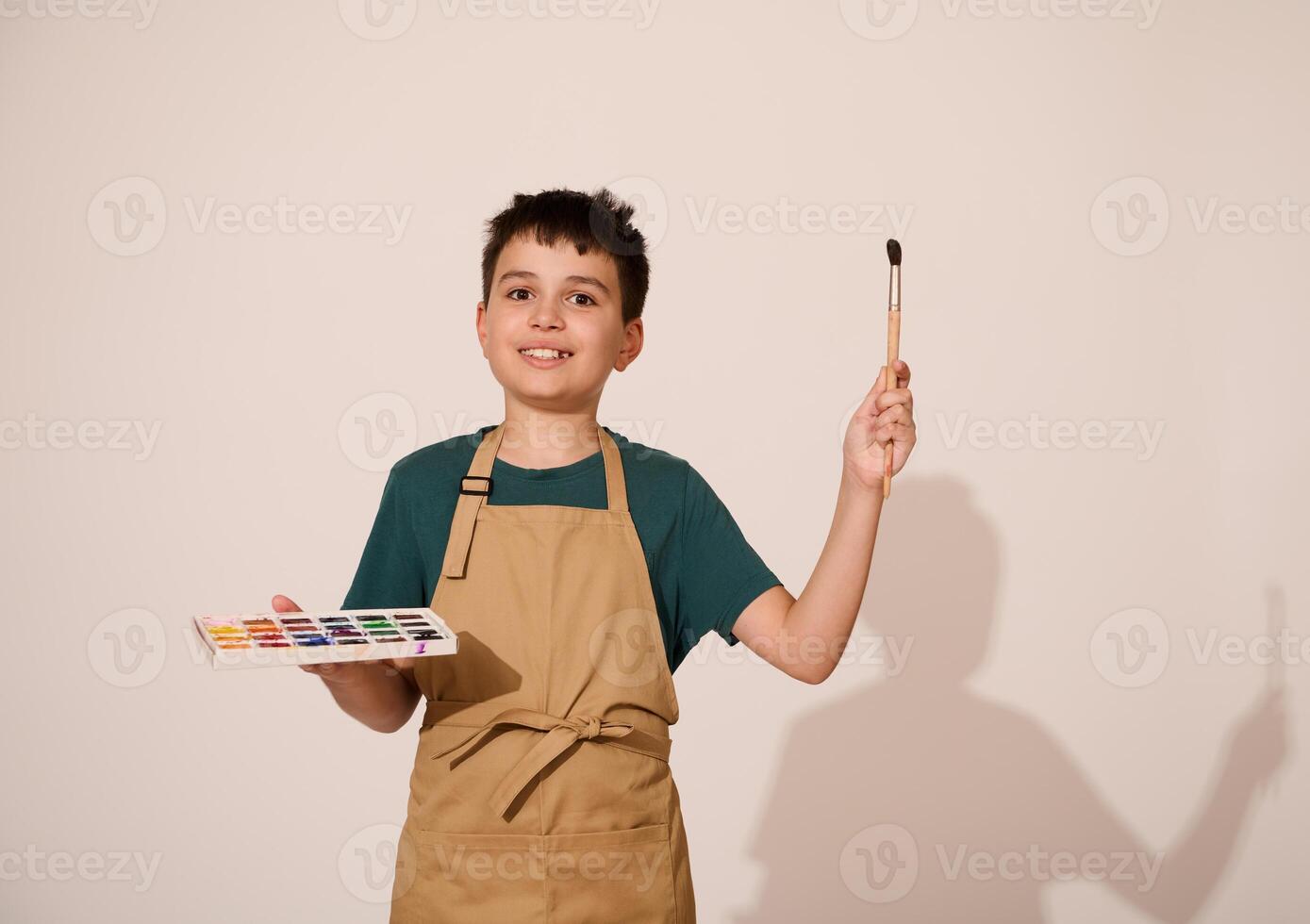 Adorable child boy enjoying art class, points a brush aside a copy space, holding a palette of vibrant watercolor paints photo