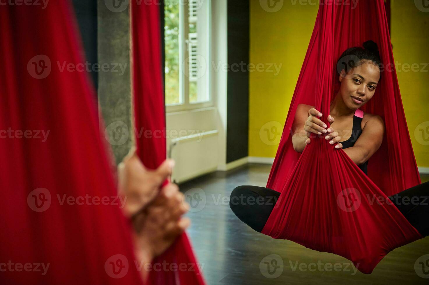 Reflection on the mirror of a sporty African pretty woman with slim body hanging on a red hammock, relaxing during aerial fly yoga stretching exercises photo