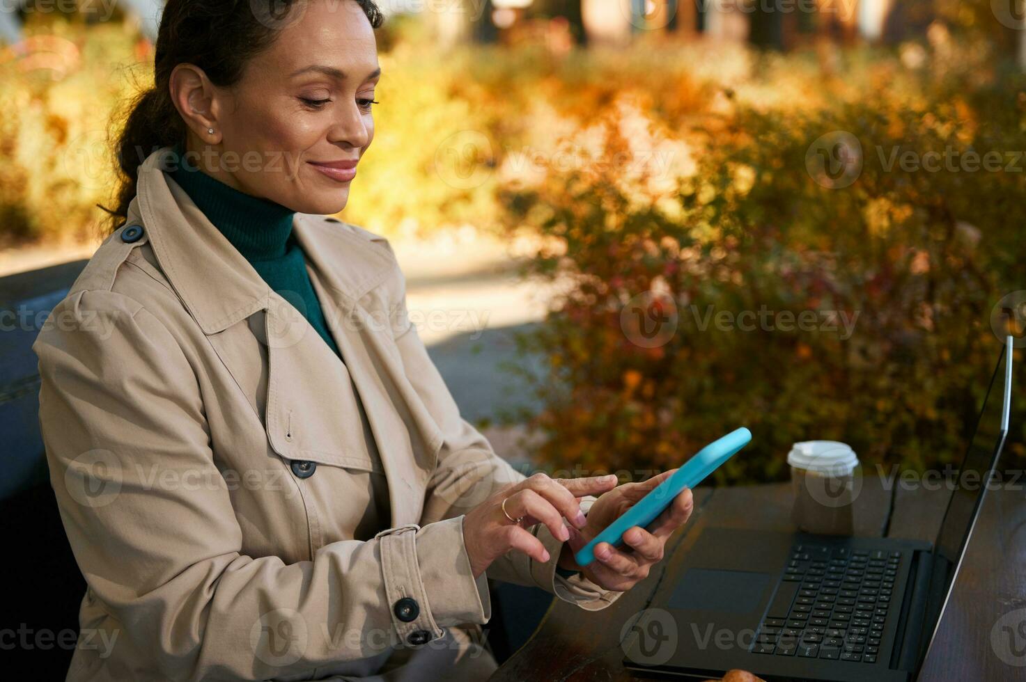 Confident dark-haired beautiful woman cutely smiles, uses mobile phone while working distantly on an oak grove wooden cafe in a beautiful autumn warm sunny day. Autumn background and business concept photo