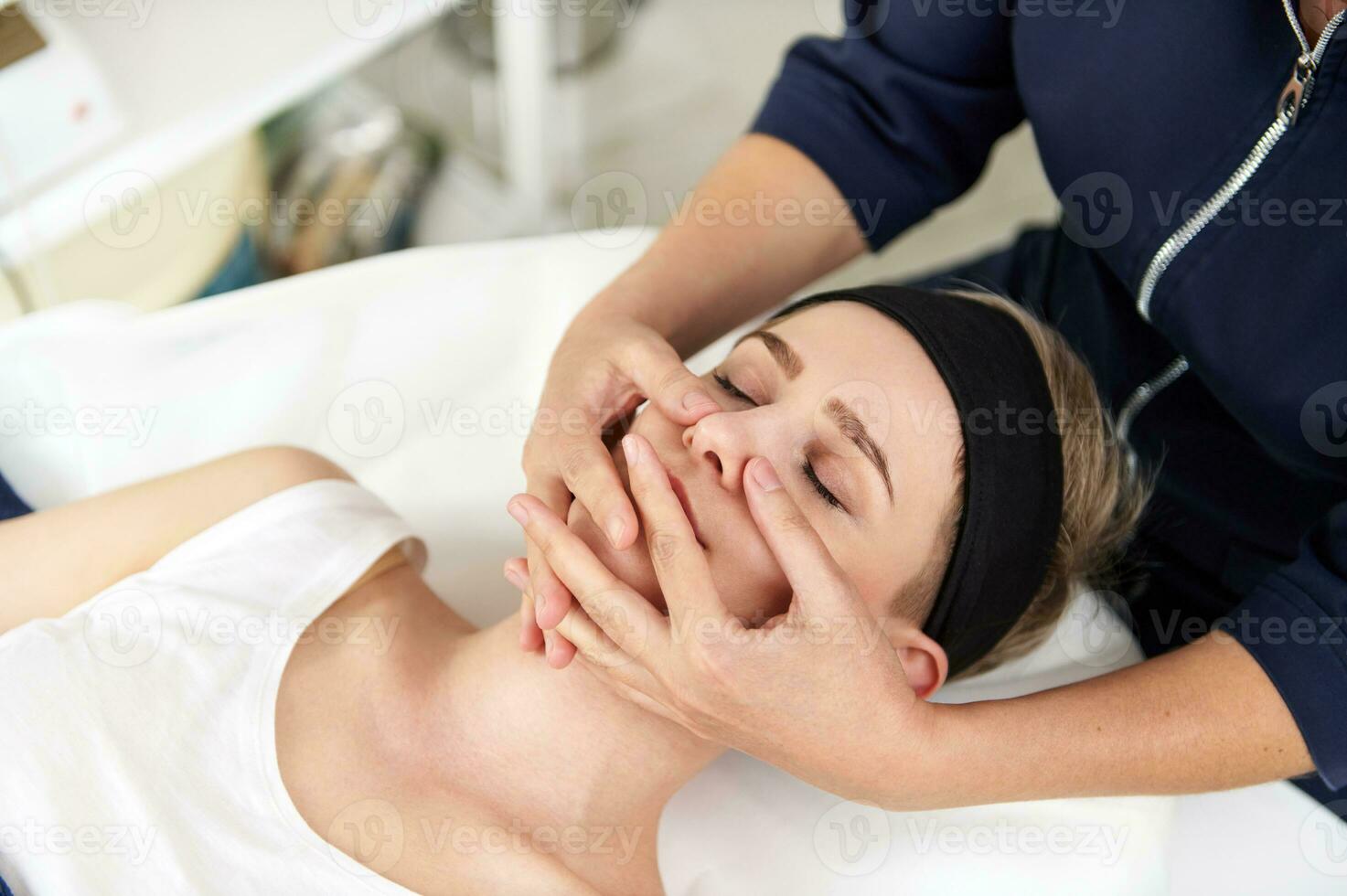 Close up portrait of aesthetician hands doing professional rejuvenating face massage to young blonde woman relaxing on massage table at spa salon photo