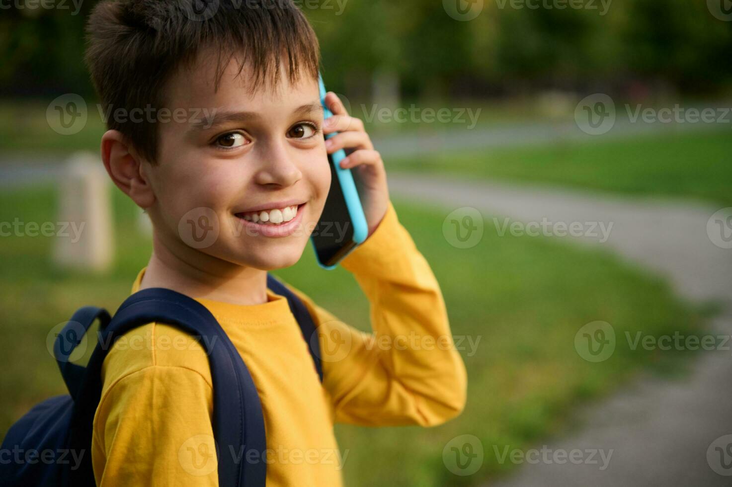 de cerca retrato de un alegre simpático adorable colegial, en amarillo camisa de entrenamiento hablando en móvil teléfono, sonriente con con dientes sonrisa mirando a cámara en el ciudad parque antecedentes foto