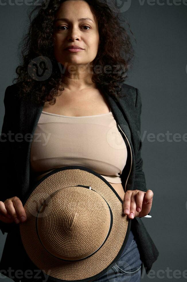 Authentic confident adult gravid woman in stylish blazer, looks at camera, holds a straw hat, hiding her pregnant belly photo