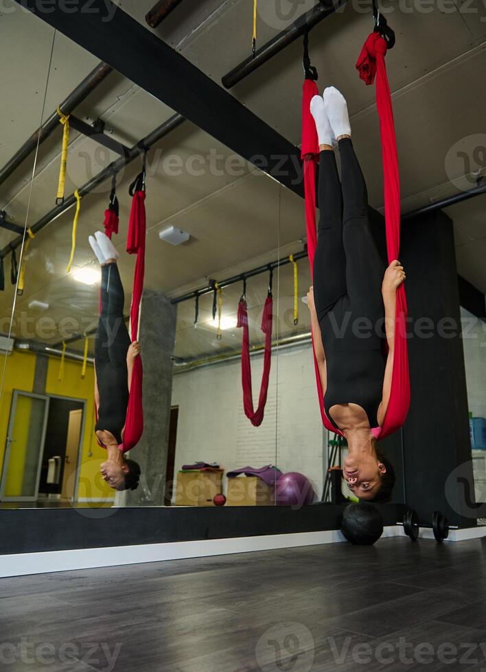 encantador juguetón joven africano americano ajuste mujer practicando aéreo volador yoga, haciendo extensión ejercicios colgando al revés abajo en rojo hamaca en yoga estudio foto