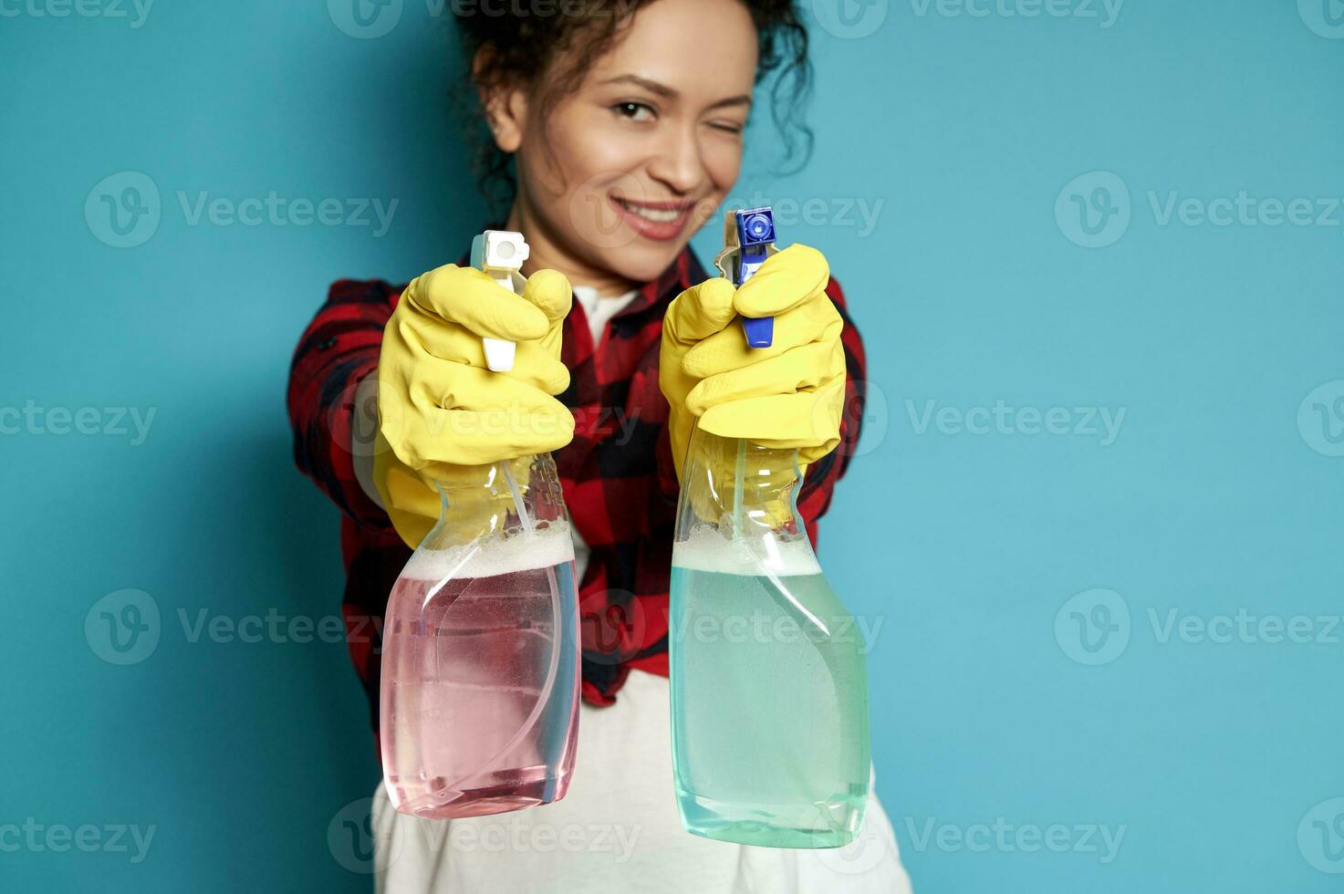 Beautiful young woman, housewife directs cleaning sprays as if shooting from a pistol, smiling maliciously and covering one eye at the camera. Focus on hands photo