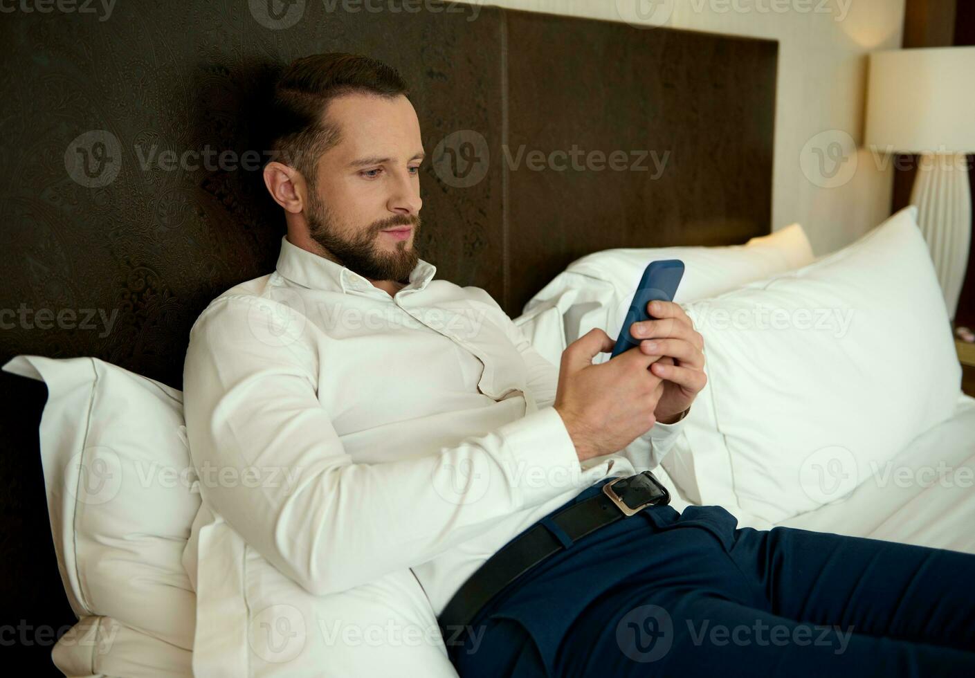 Handsome young Caucasian bearded man, successful businessman browsing and chatting on mobile phone, lying on a bed in the bedchamber of a hotel, relaxing after hard day at work and business meetings photo
