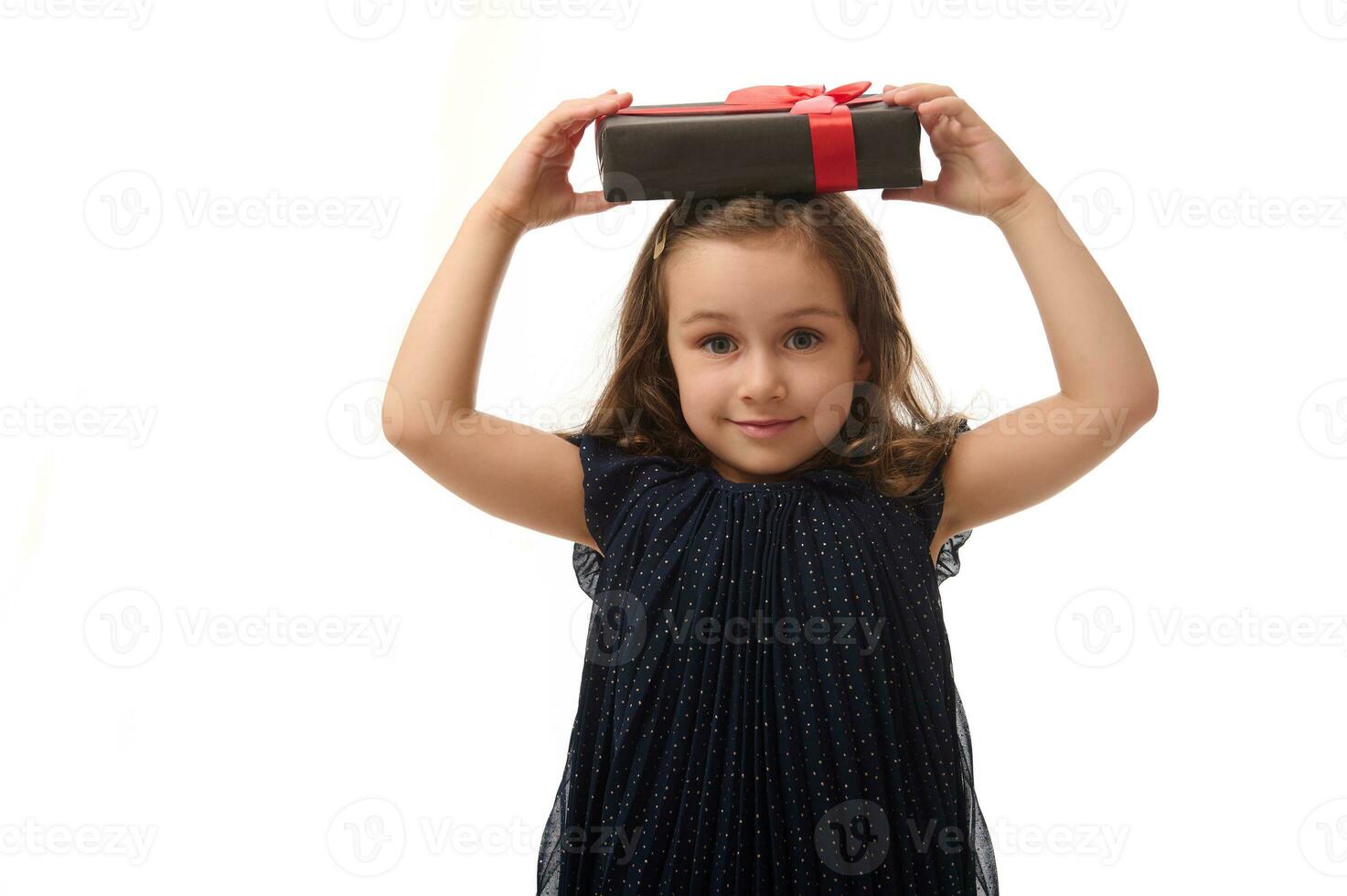 atractivo sonriente pequeño niña en noche vestir participación un negro regalo caja con rojo cinta en su cabeza, mirando a cámara. aniversario, negro viernes concepto. aislado en blanco fondo, Copiar espacio foto