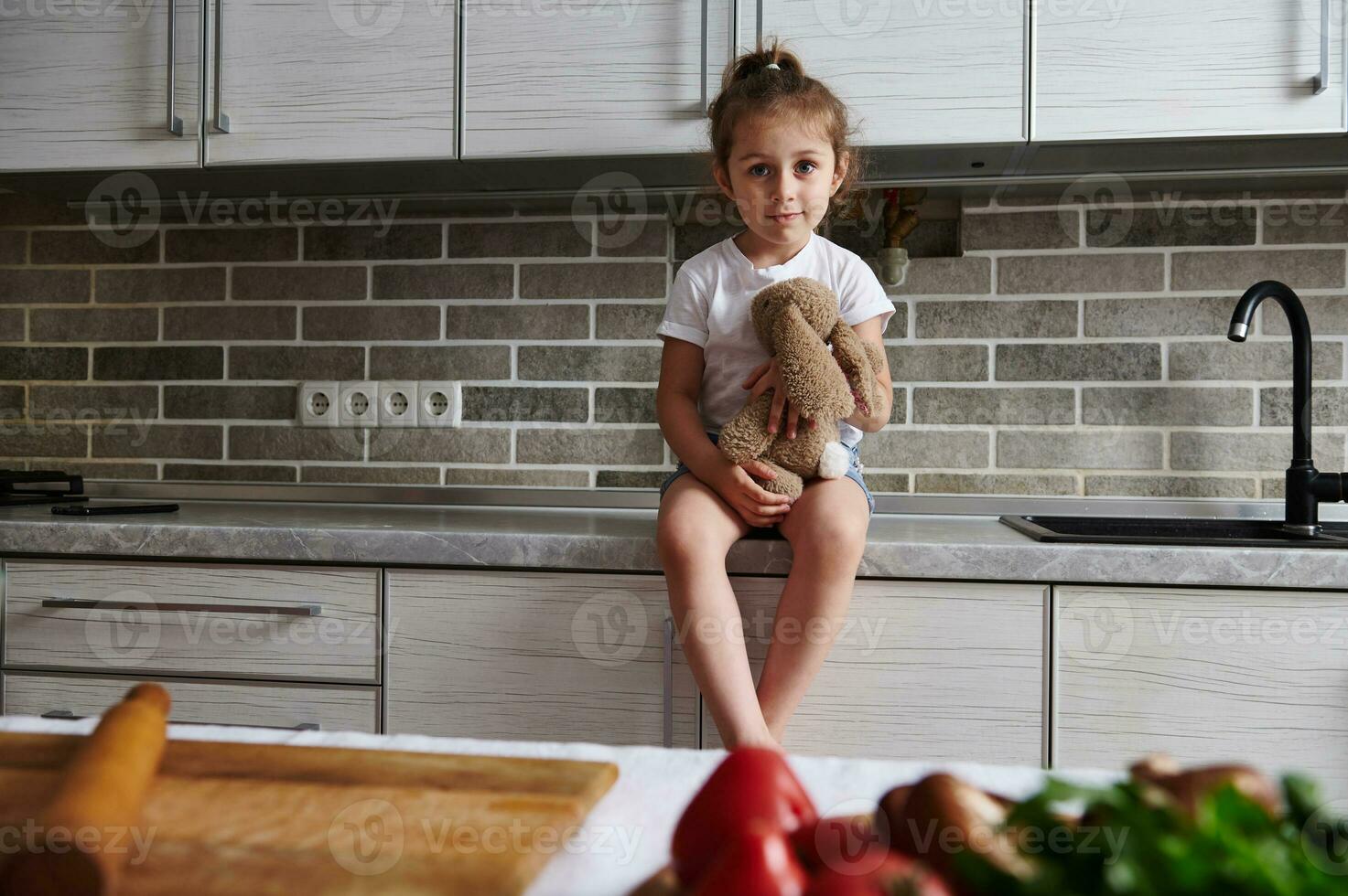 A charming little girl is sitting on a kitchen countertop with a soft plush toy in her hands. Vegetables on the table in the foreground photo