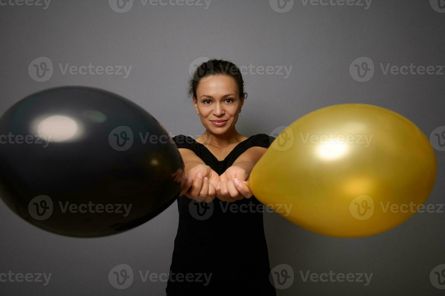 Beautiful woman dressed in black smiles looking at camera and poses against gray background holding gold and black air balloons in her outstretched hand. Black Friday concept with copy ad space photo