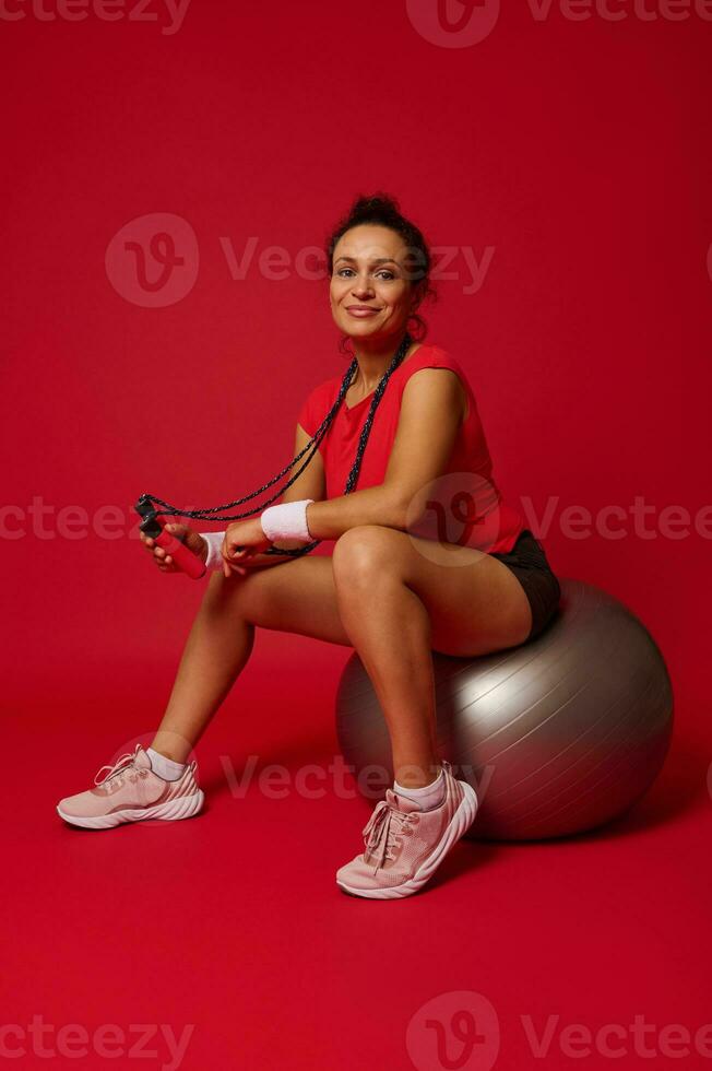 hermosa atlético mujer en rojo apretado parte superior y negro Deportes pantalones cortos participación saltar cuerda y sonriente a cámara mientras sentado en plata ajuste pelota en rojo antecedentes con Copiar espacio foto