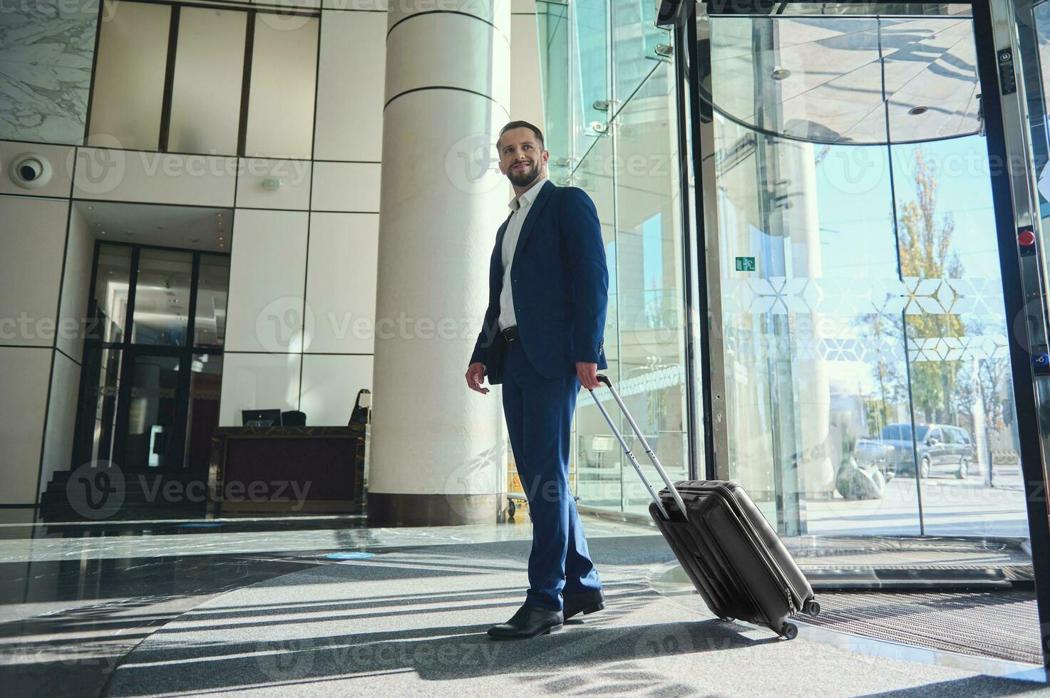 A full-length handsome businessman developer entrepreneur in a business suit with a suitcase stands at the entrance of a modern luxury building, walking to the lobby during his business trip photo