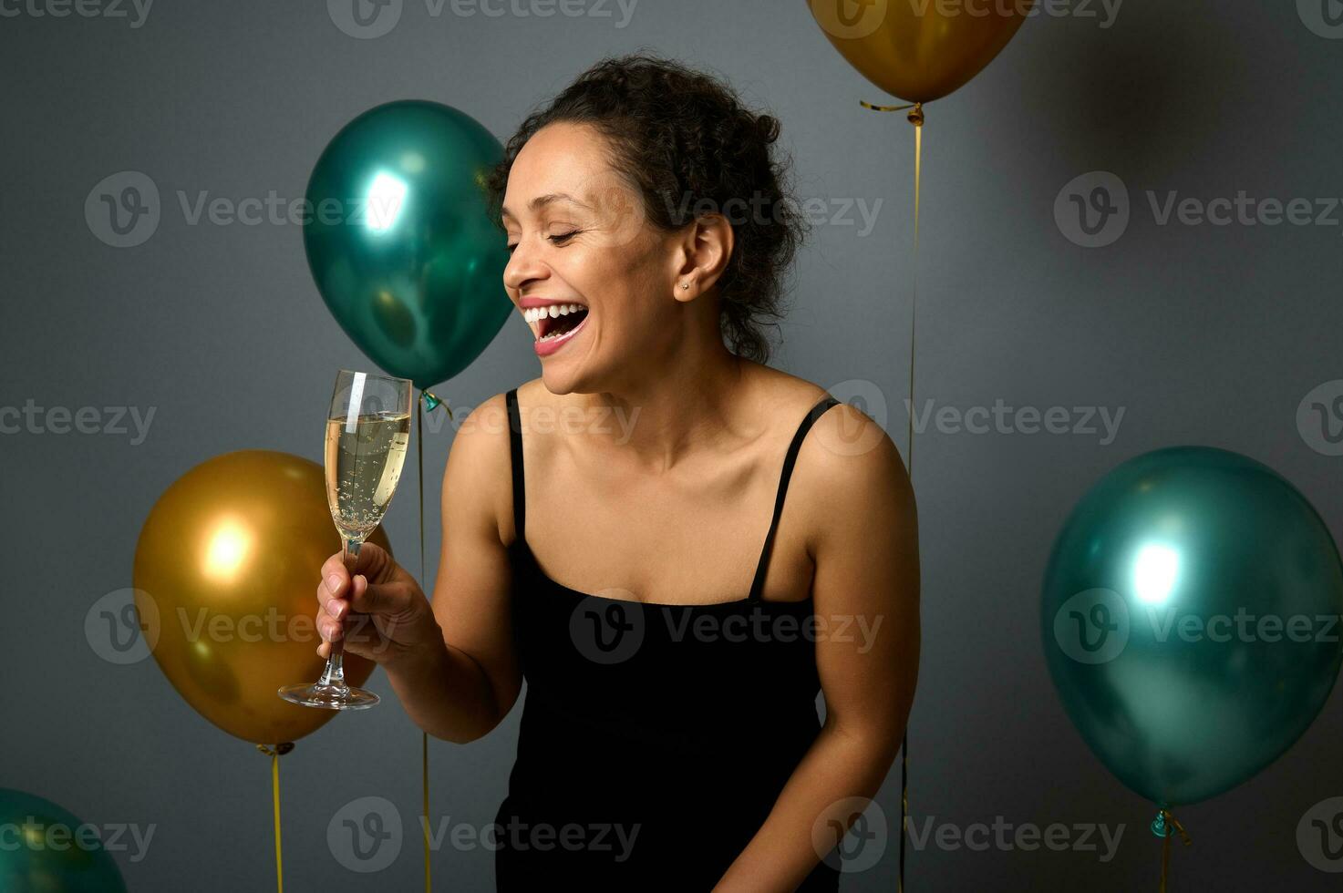 Happy African American cute woman in black evening dress laughs, rejoices, enjoys birthday party, poses over gray background with beautiful air balloons. Christmas, New Years and events concept for ad photo