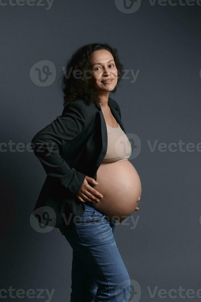 alegre joven embarazada mujer en azul vaqueros, gris chaqueta de sport y beige sostén, participación su barriga, sonriente mirando a cámara foto
