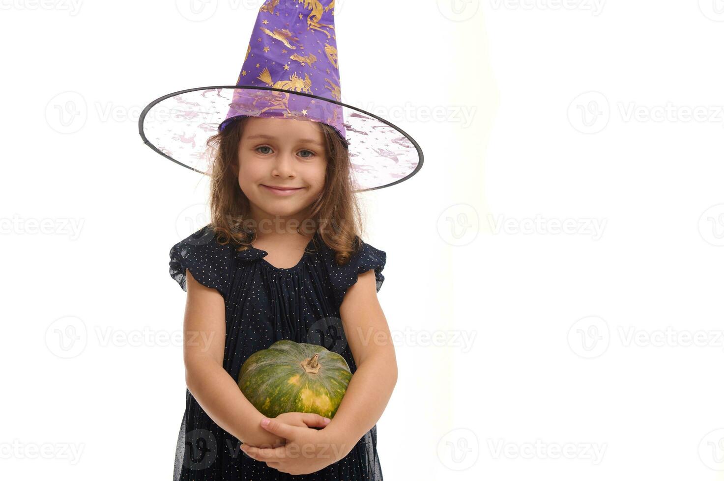 Halloween Witch concept - Portrait of little Caucasian girl dressed in stylish carnival attire and wizard hat, witch child smiles posing with pumpkin against a white background with copy space photo