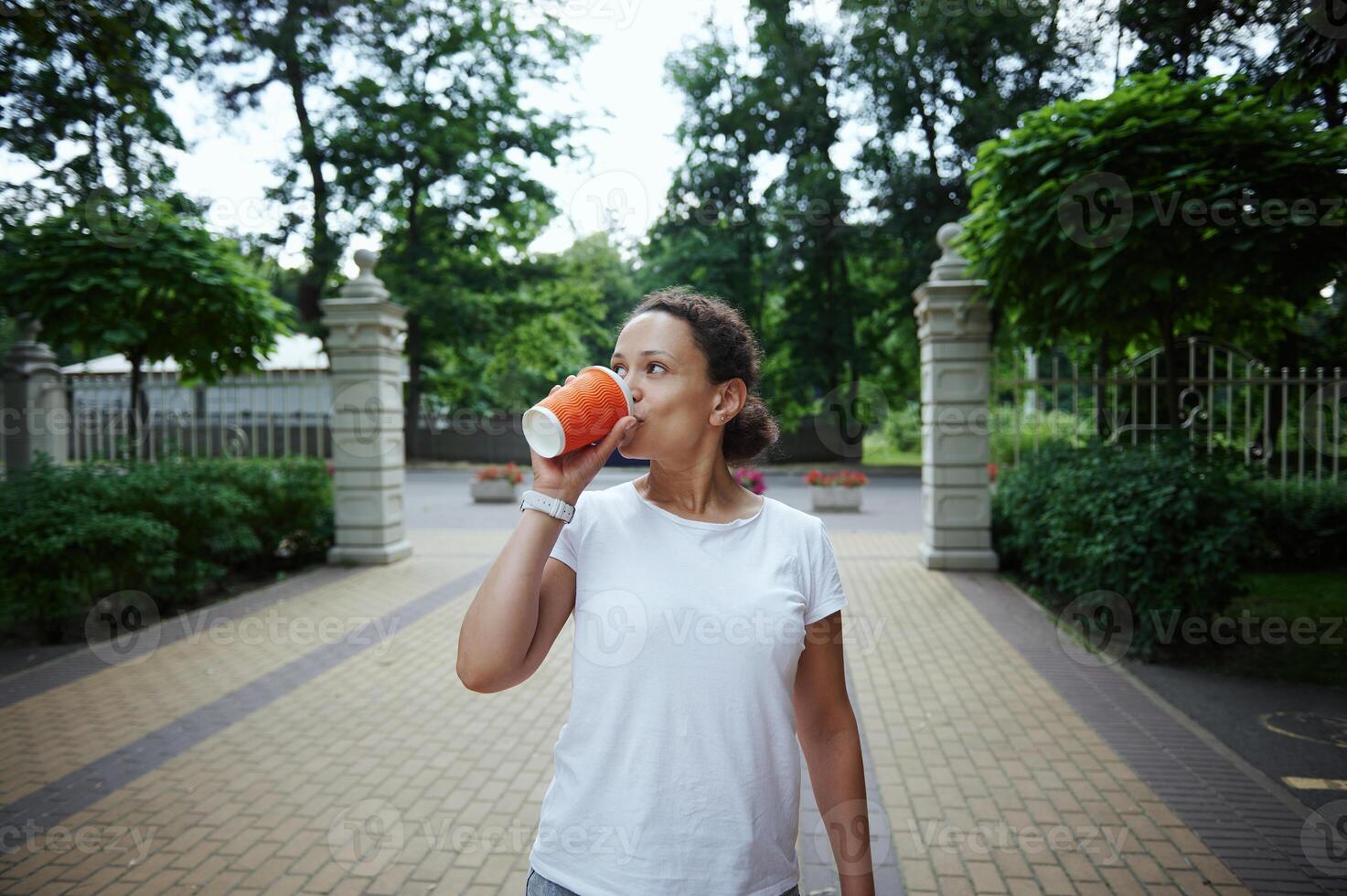 Charming young woman drinking takeaway coffee from a disposable cardboard mug, while enjoying her walk in park photo