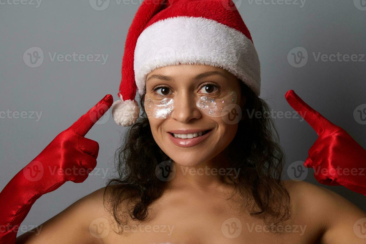 Attractive brunette in Santa costume points fingers on eye patches on her face, smiles looking at camera. Advertising for beauty salons for Christmas and New year giveaways. Spa, skin care concept photo