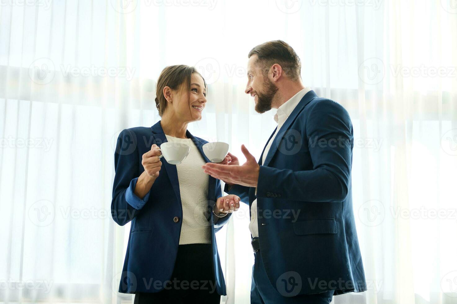 encantador mujer y hermoso hombre en Armada negocio trajes teniendo un conversacion terminado un taza de café durante un trabajo romper, que se discute planes y objetivos, en pie por el ventana y sonriente a cada otro foto